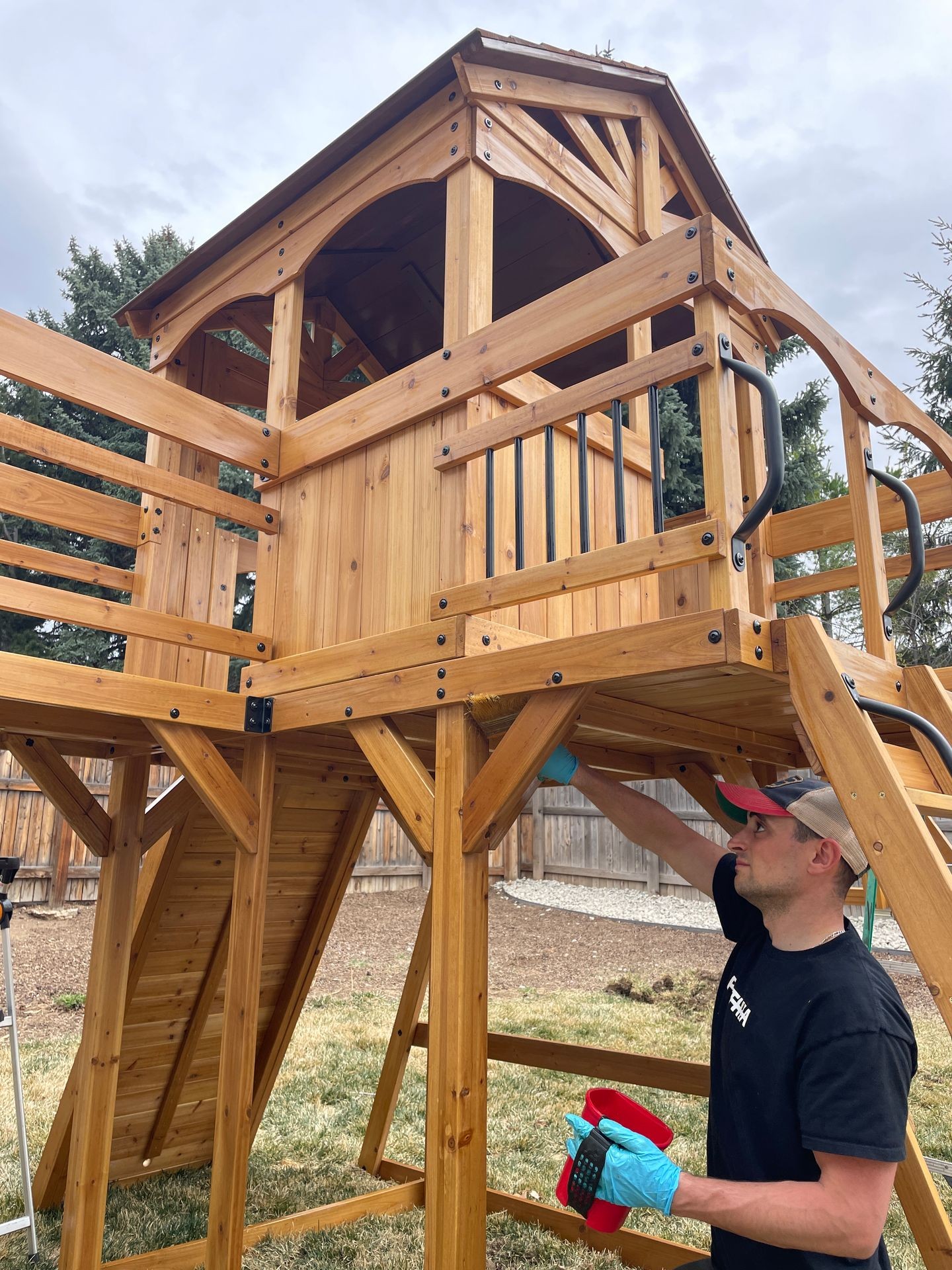 Person applying stain to a wooden playhouse in a backyard, wearing gloves and holding paint supplies.