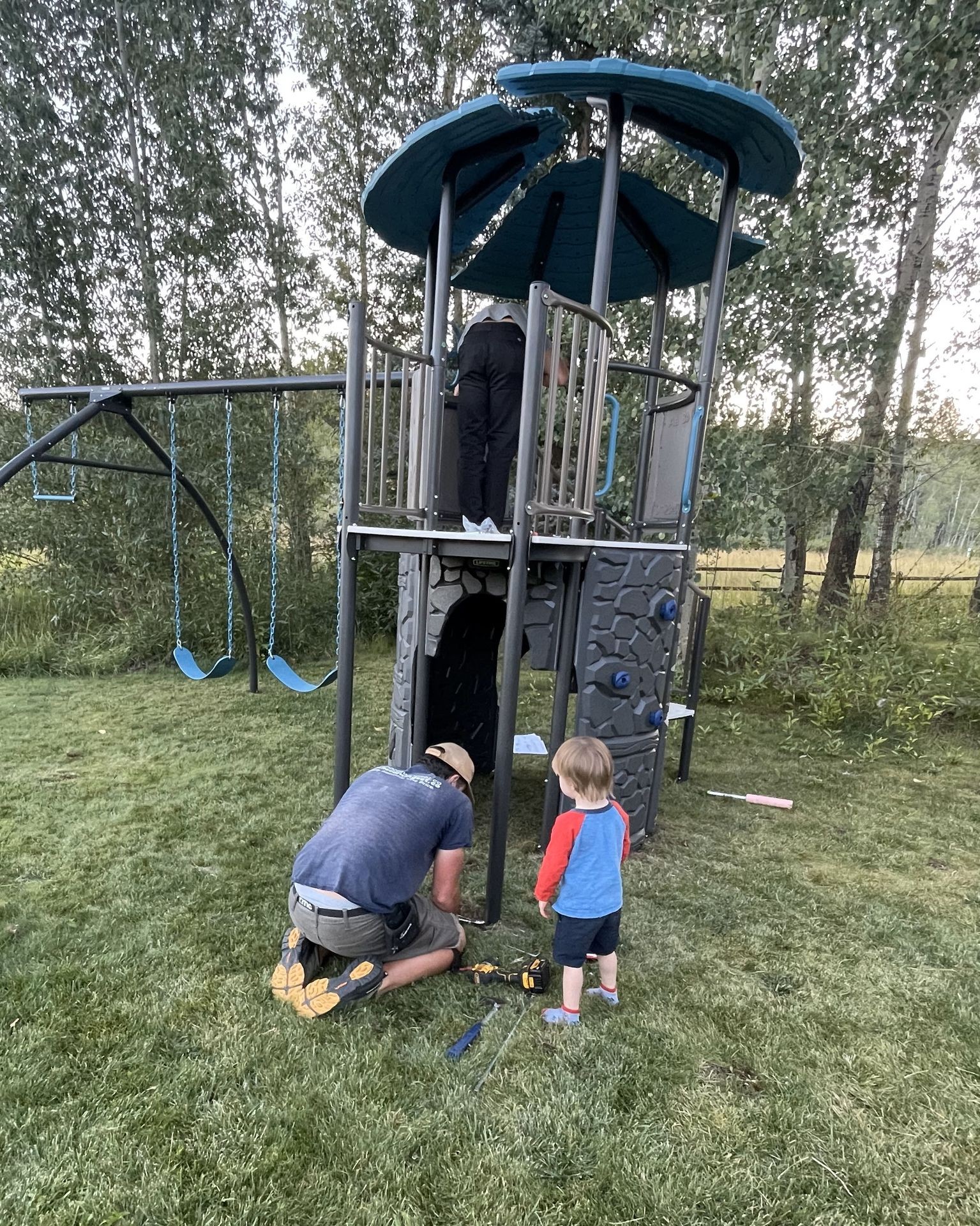 Child and adult assembling a playground slide outdoors in a grassy area surrounded by trees.